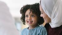 A toddler boys sits on his mother's lap at the doctor's office. They are sitting at a table and a male doctor is sitting across from them. The mother and child are smiling at the doctor as he speaks.